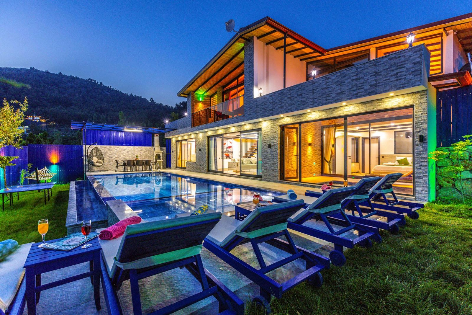 Timber-and-glass villa glowing at dusk beside a pool with loungers